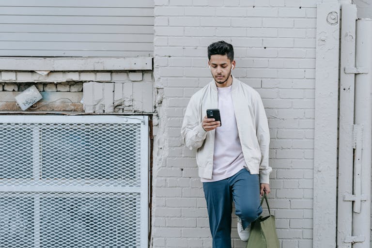 Stylish ethnic male with eco bag listening to song from wireless earbuds while text messaging on cellphone against brick wall