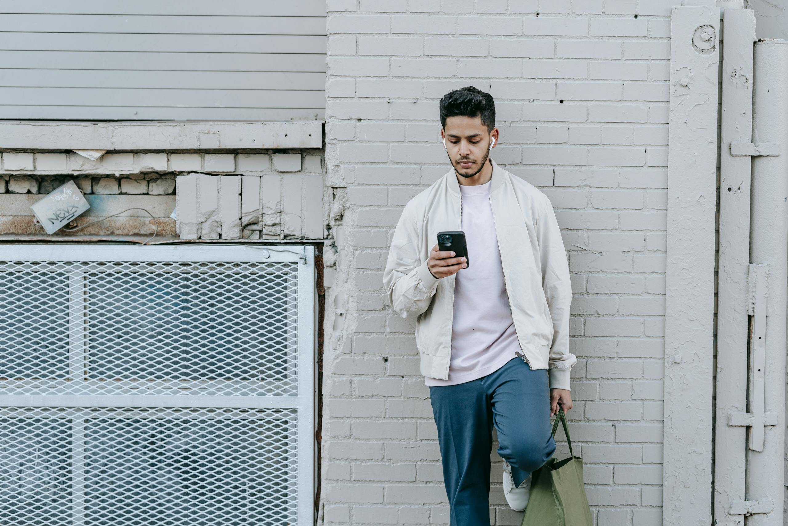 Stylish ethnic male with eco bag listening to song from wireless earbuds while text messaging on cellphone against brick wall