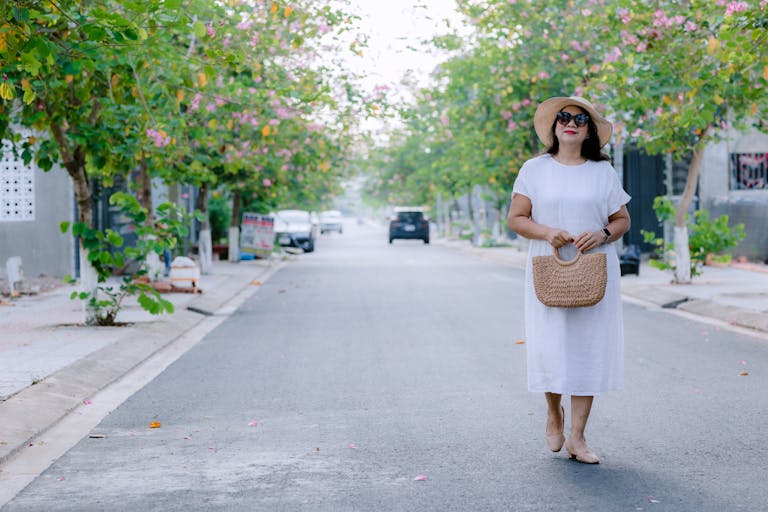Stylish woman in white dress walking on a tree-lined street with blooming flowers.