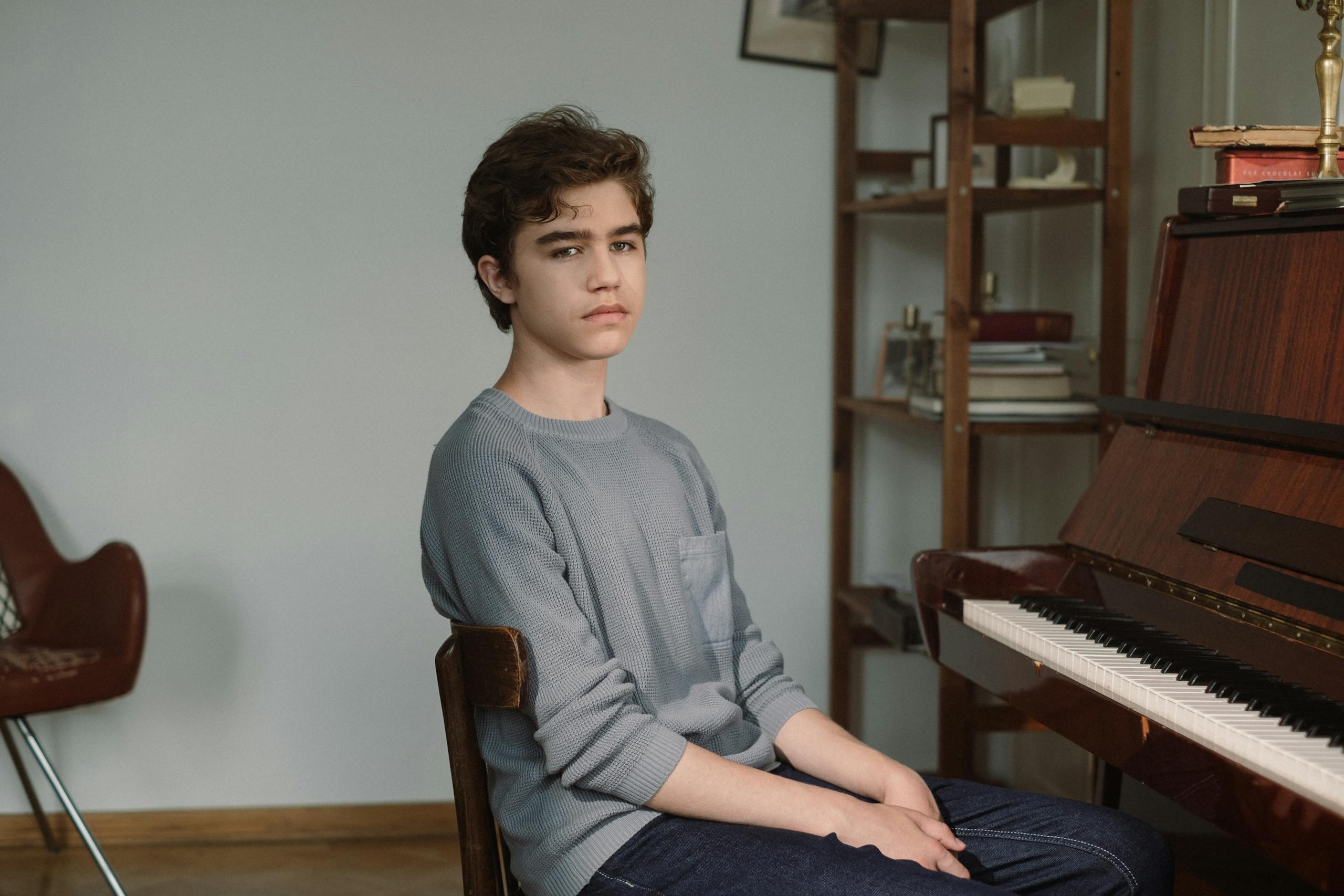 Teen boy seated by piano in a cozy room, wearing a sweater. Musical ambiance.