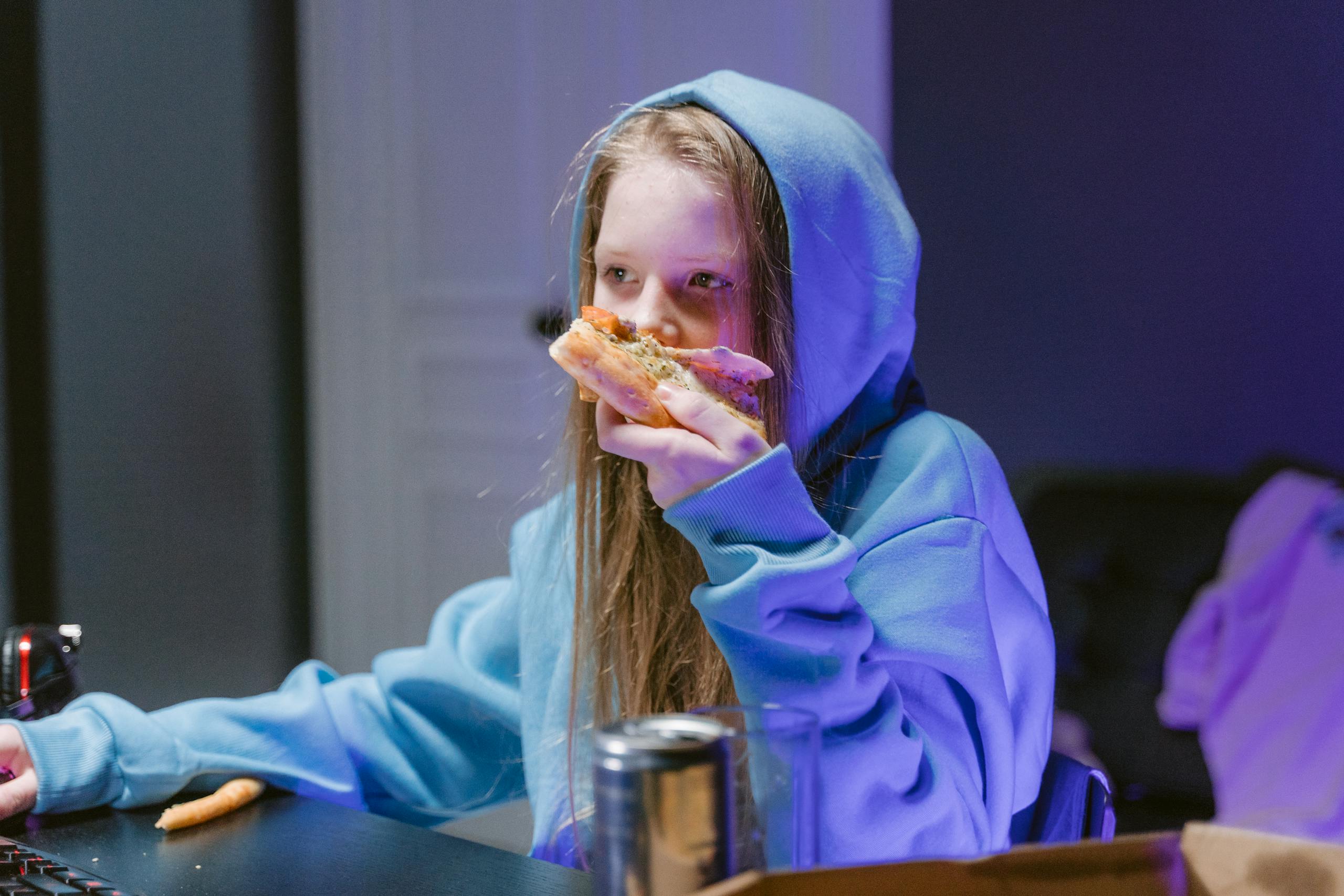 Teen girl in a hoodie enjoying pizza while gaming indoors, illuminated by screen light.