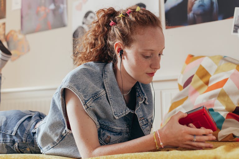 Teenage girl in denim vest listening to music on a retro cassette player while lying on bed.