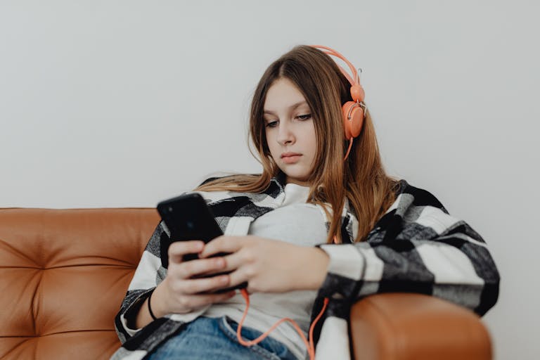 Teenage girl sitting on a sofa listening to music on headphones and using a smartphone.