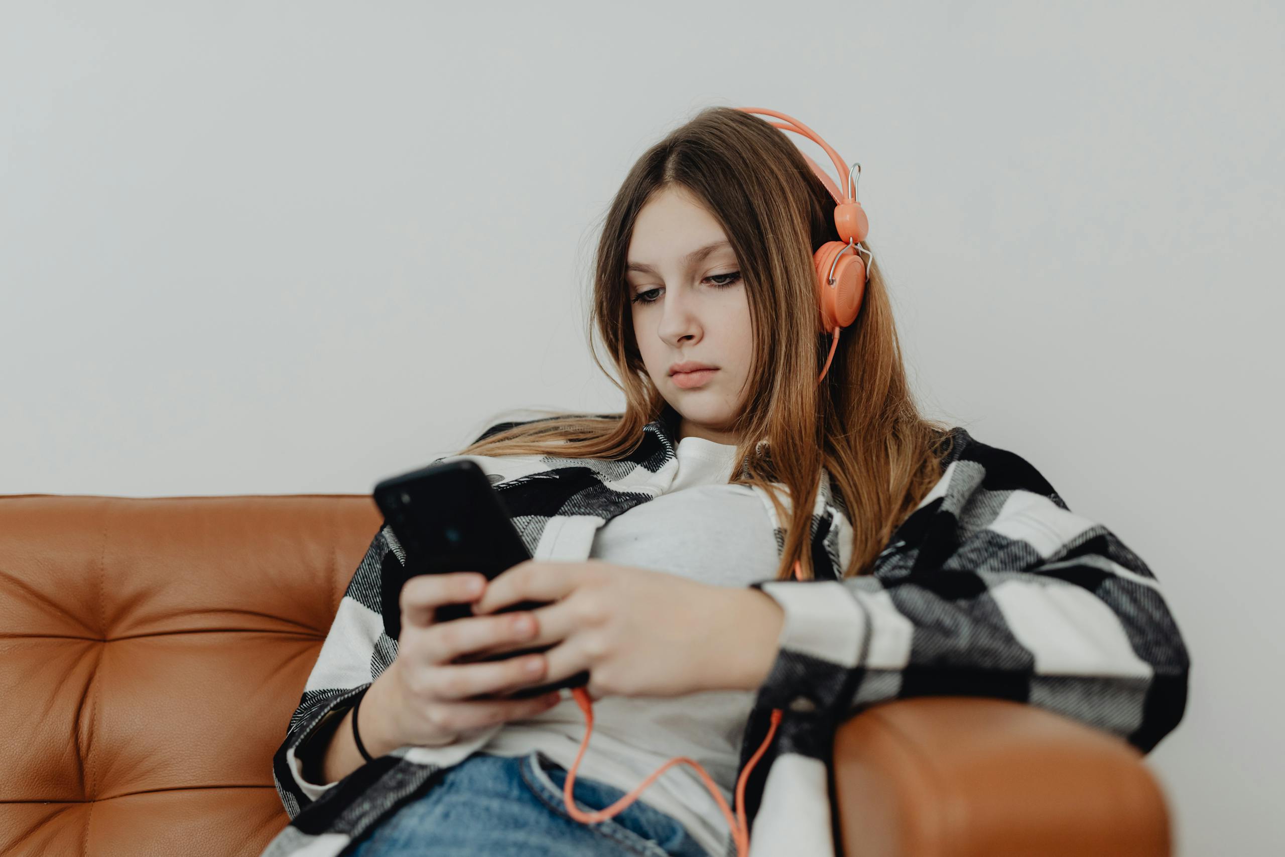 Teenage girl sitting on a sofa listening to music on headphones and using a smartphone.