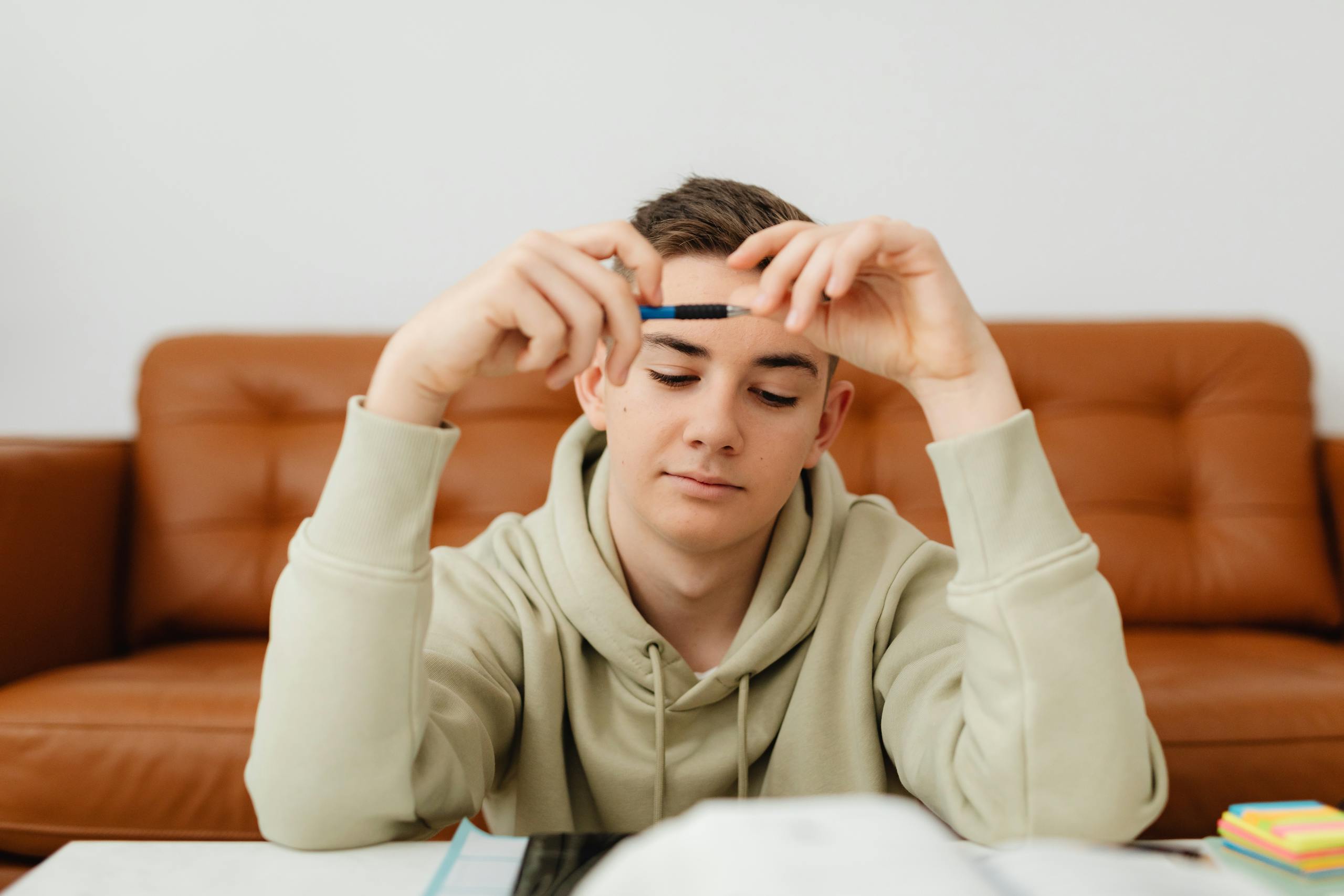 Teenager in a hoodie focuses while studying indoors, holding a pen. Bright and thoughtful atmosphere.