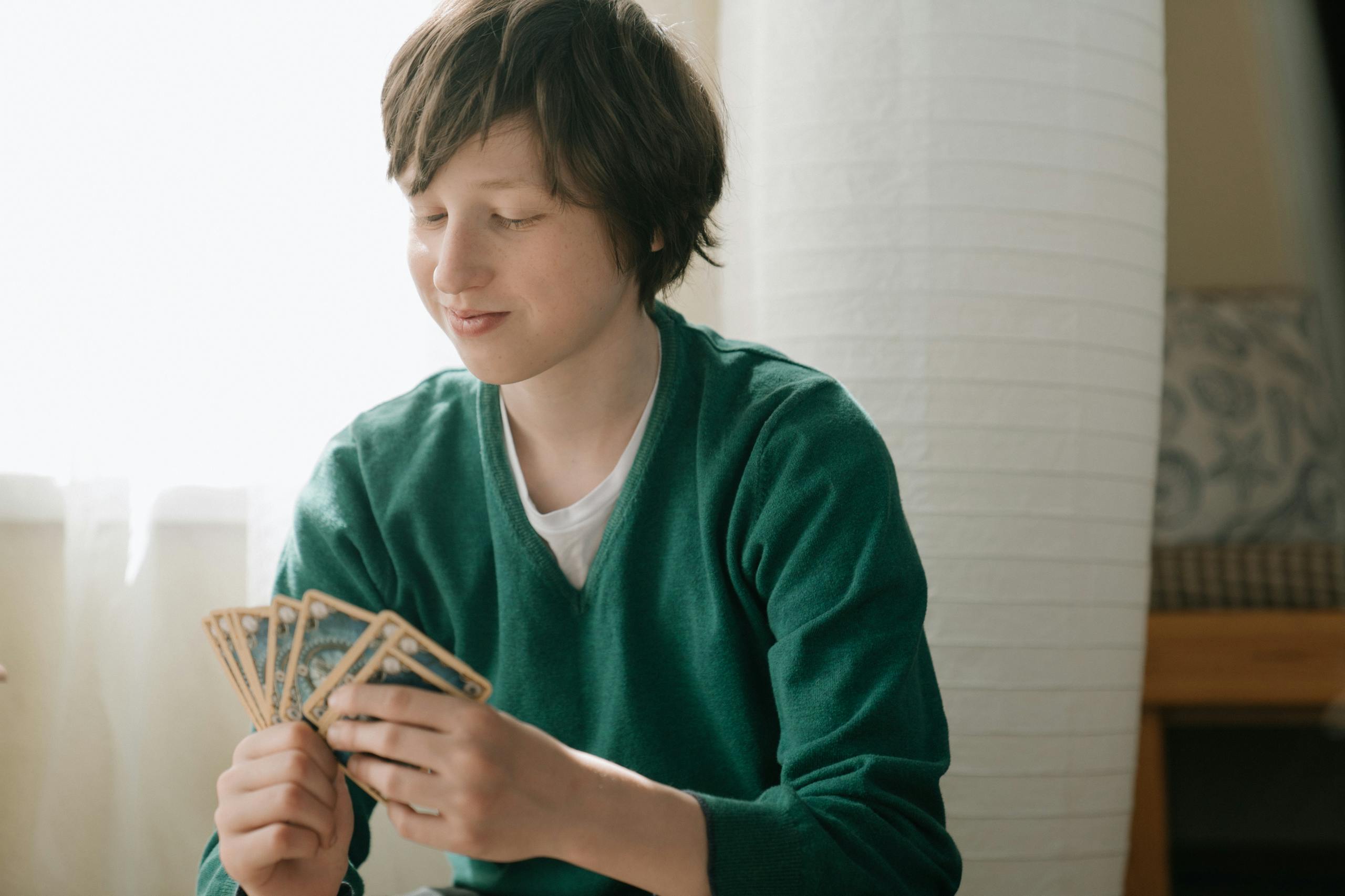 Teenager in green sweater smiling while playing a strategic card game indoors.