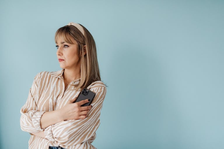 Thoughtful woman in striped shirt holding a smartphone against a light blue background.