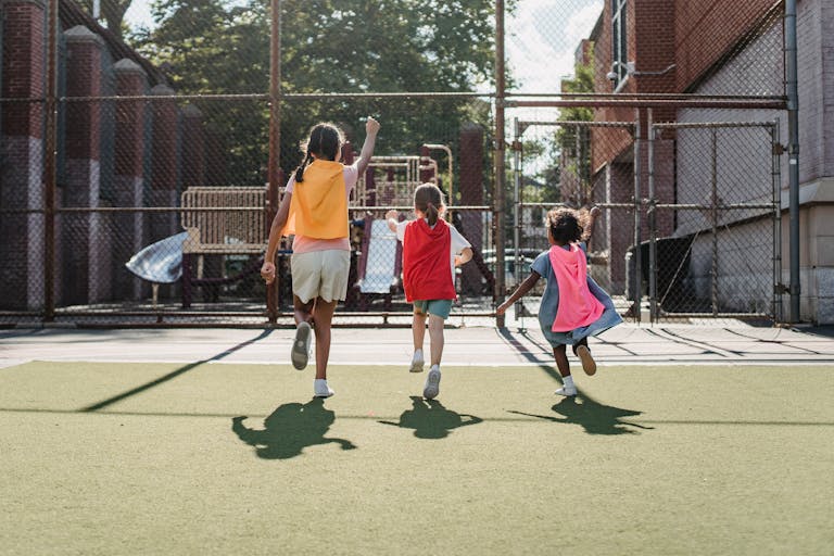 Three children playing as superheroes on a sunny playground, wearing colorful capes.