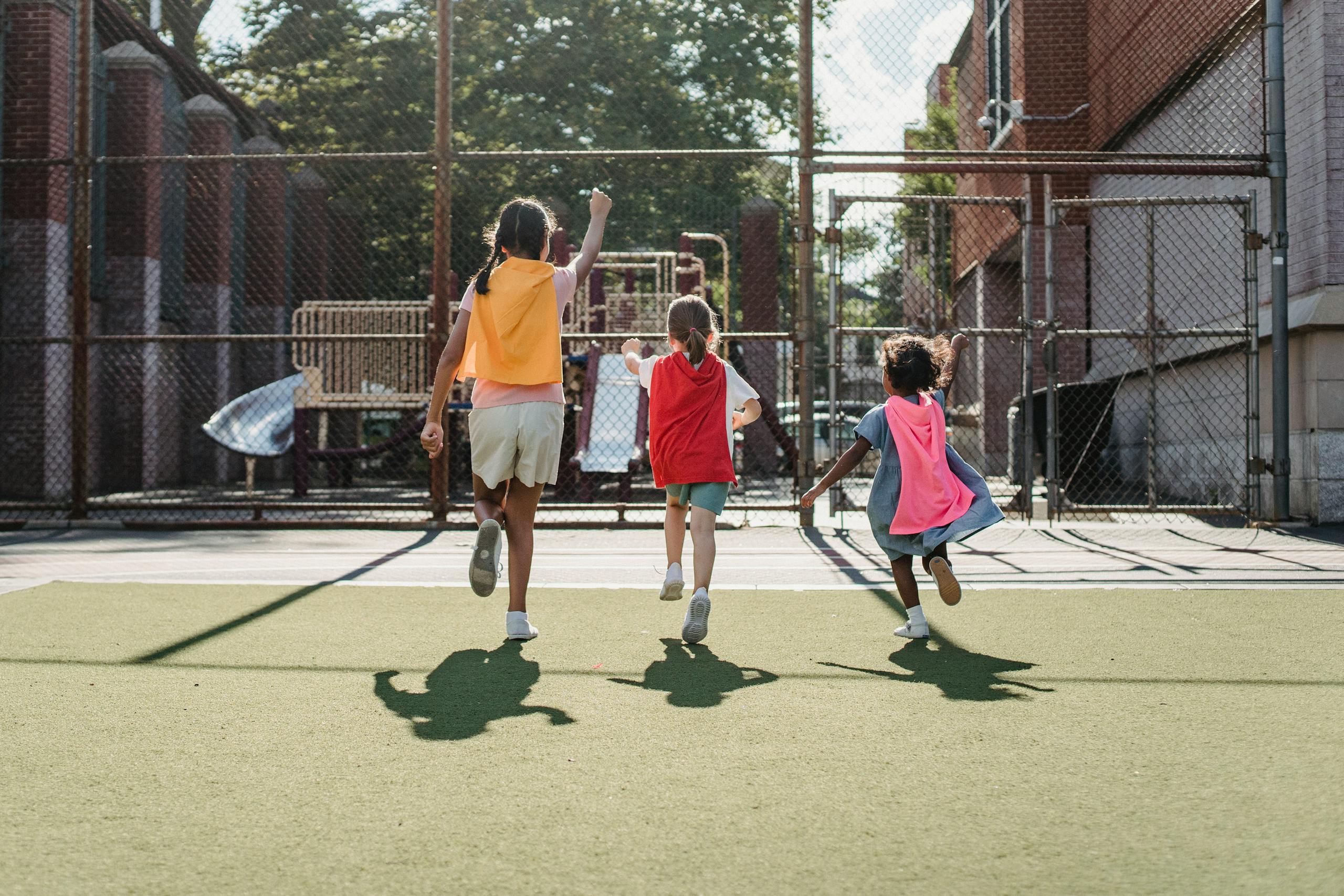 Three children playing as superheroes on a sunny playground, wearing colorful capes.