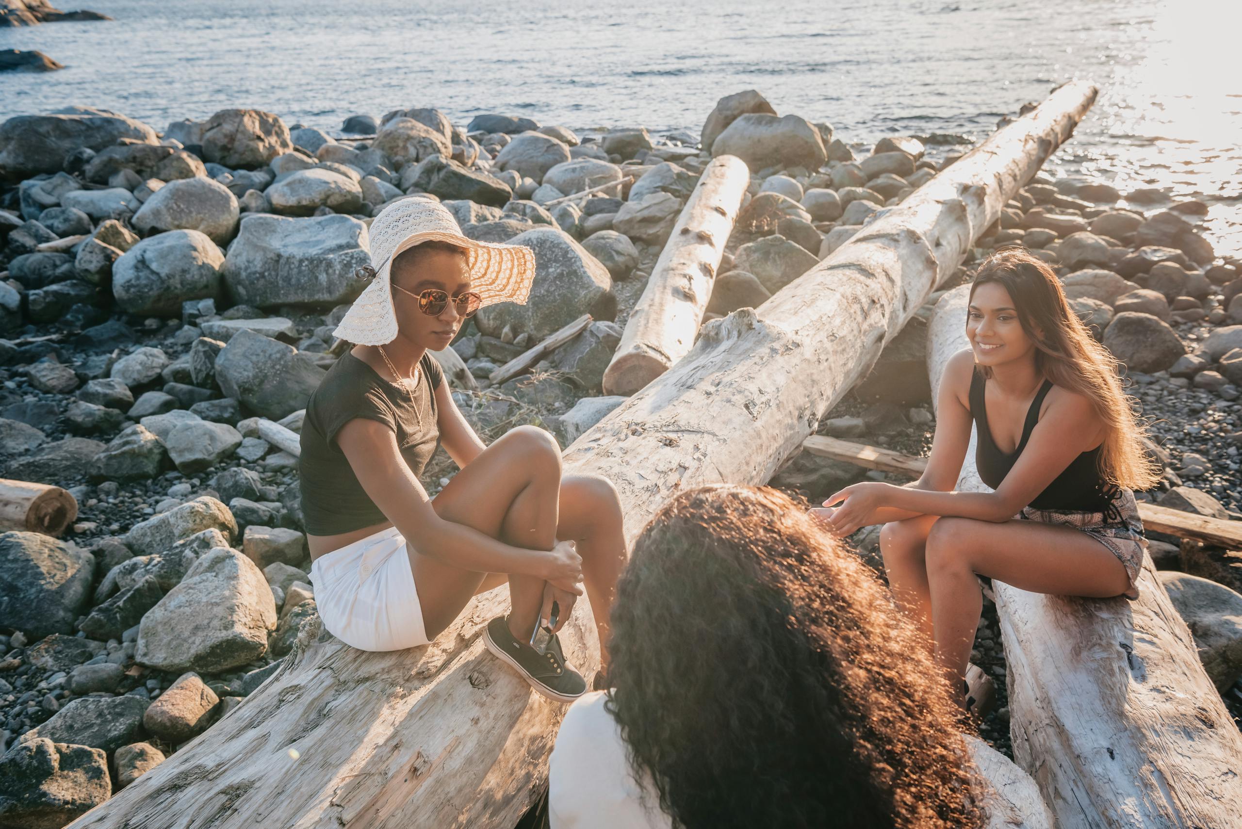 Three women enjoy a relaxing conversation on a rocky beach, sitting on large logs by the sea.