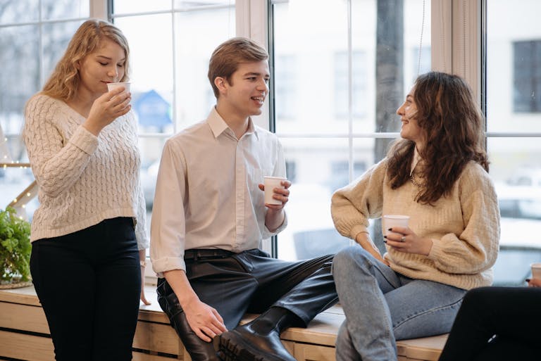Three young adults enjoying coffee and conversation in a relaxed indoor setting by the window.