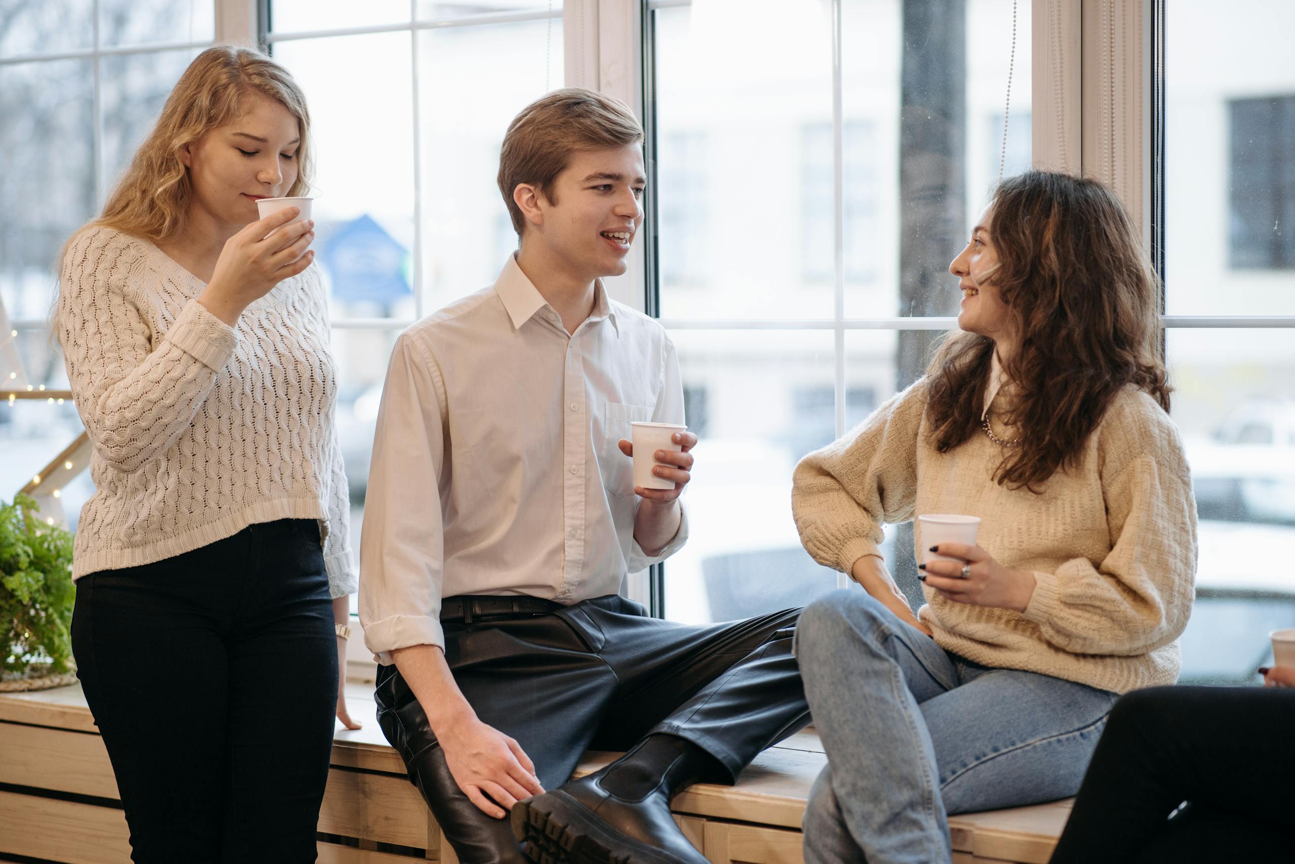 Three young adults enjoying coffee and conversation in a relaxed indoor setting by the window.