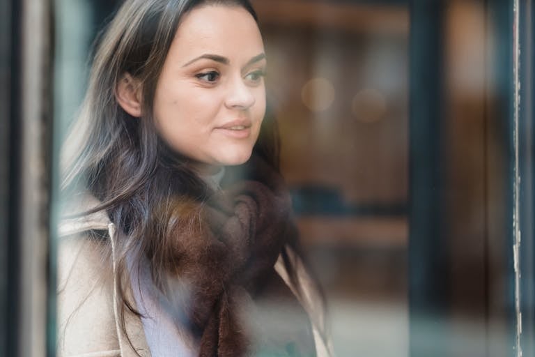 Through glass of crop positive female wearing warm outerwear standing near glass wall and looking away