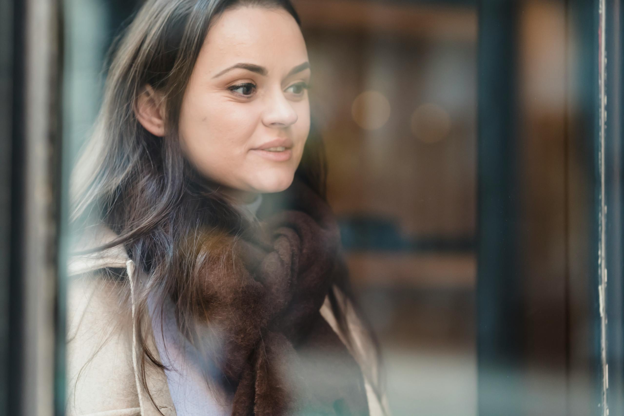 Through glass of crop positive female wearing warm outerwear standing near glass wall and looking away