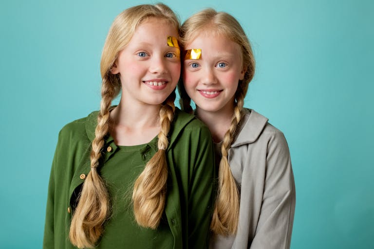 Twin sisters with blonde braids and face stickers smiling against a turquoise background.