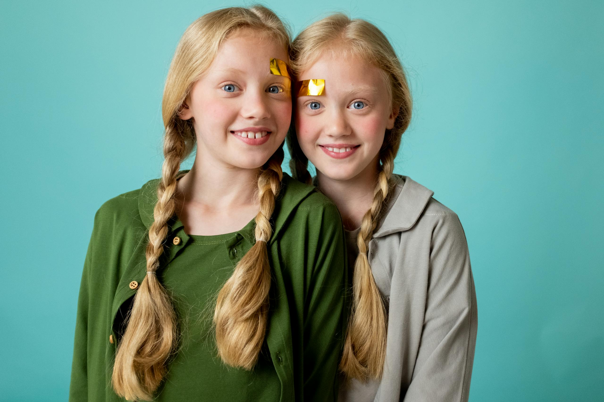 Twin sisters with blonde braids and face stickers smiling against a turquoise background.