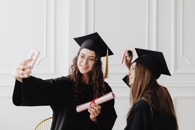 Two happy female graduates in academic gowns taking a selfie while holding diplomas indoors.