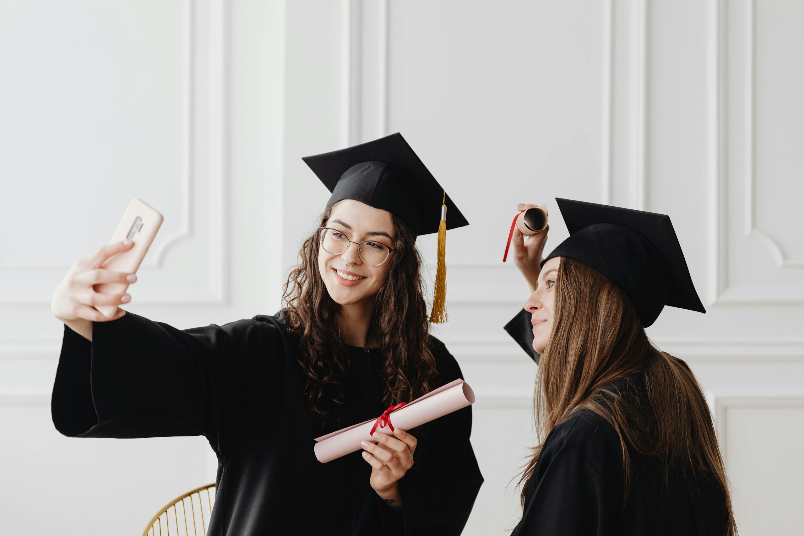 Two happy female graduates in academic gowns taking a selfie while holding diplomas indoors.