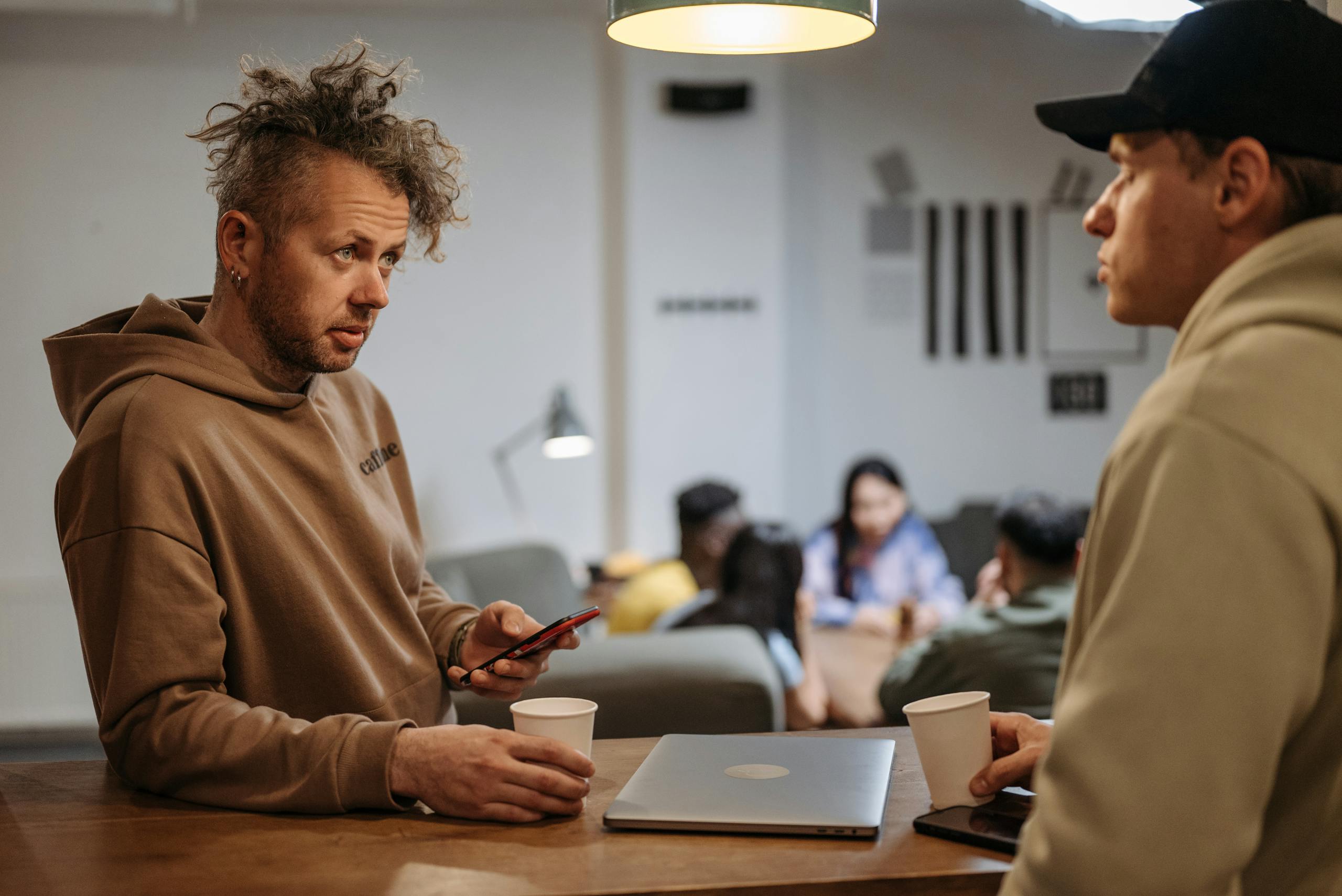 Two men in a casual office setting talking over coffee with laptops.
