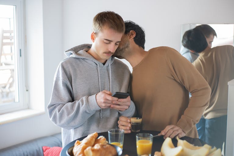 Two men in a cozy home setting enjoying breakfast, using a smartphone and sharing a moment.