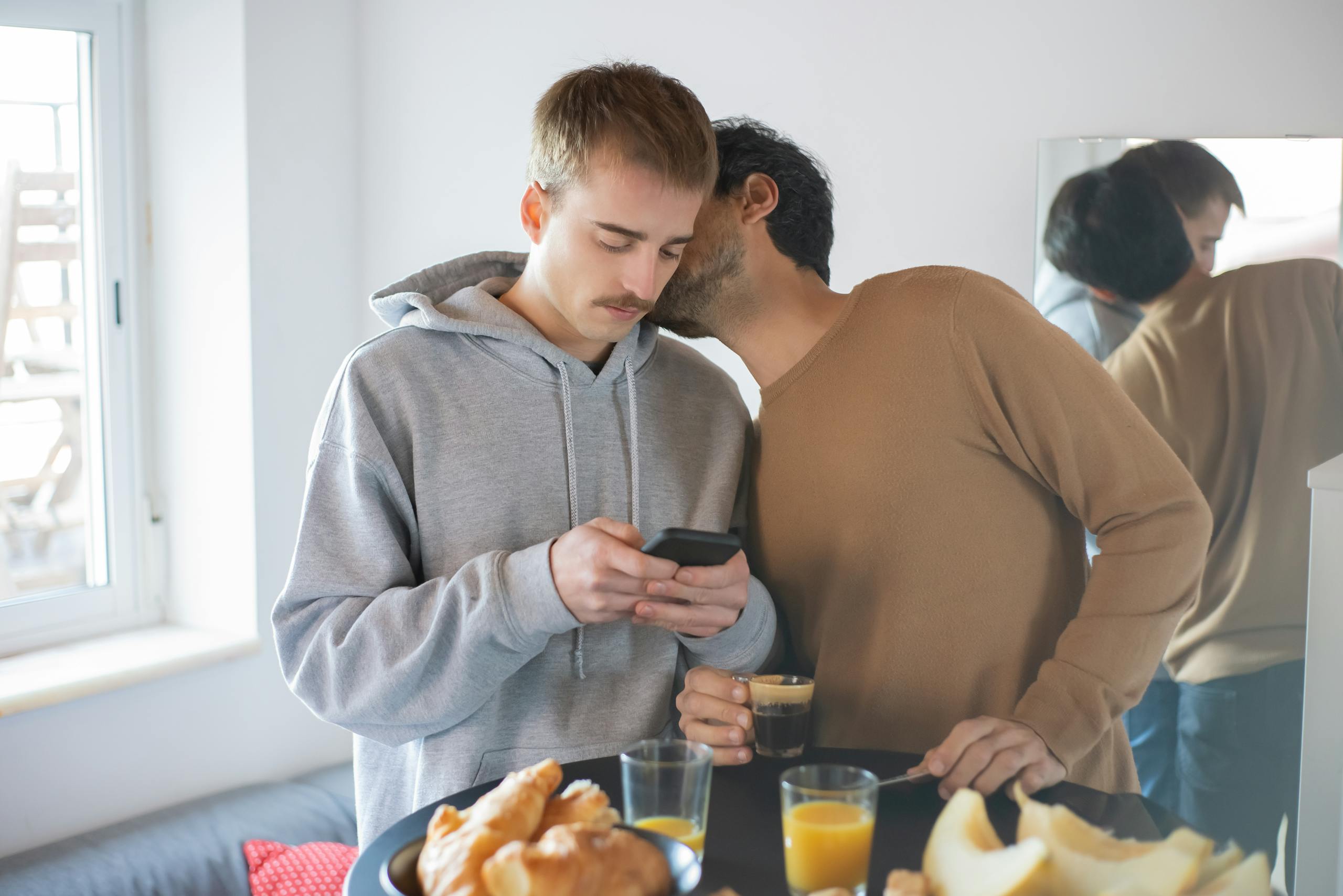 Two men in a cozy home setting enjoying breakfast, using a smartphone and sharing a moment.