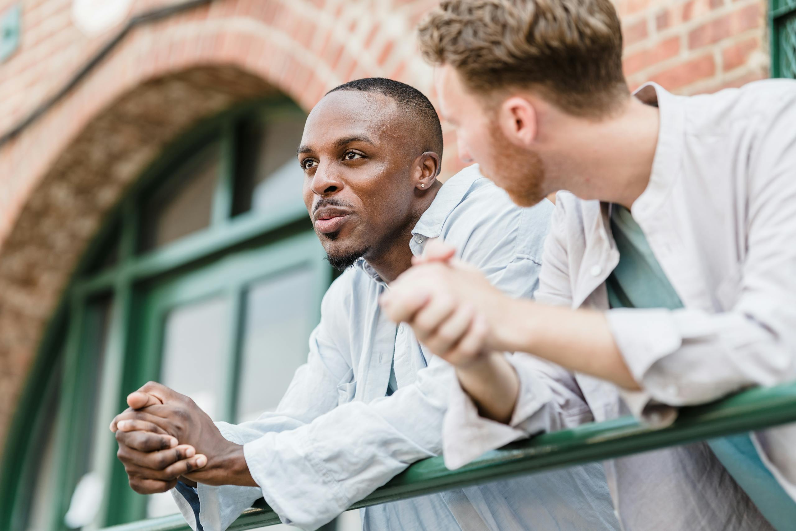Two men lean on a railing outdoors, engaged in a relaxed conversation.