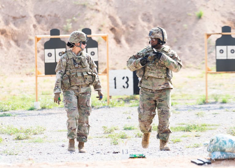 Two soldiers in military uniforms practicing at an outdoor shooting range with targets visible.