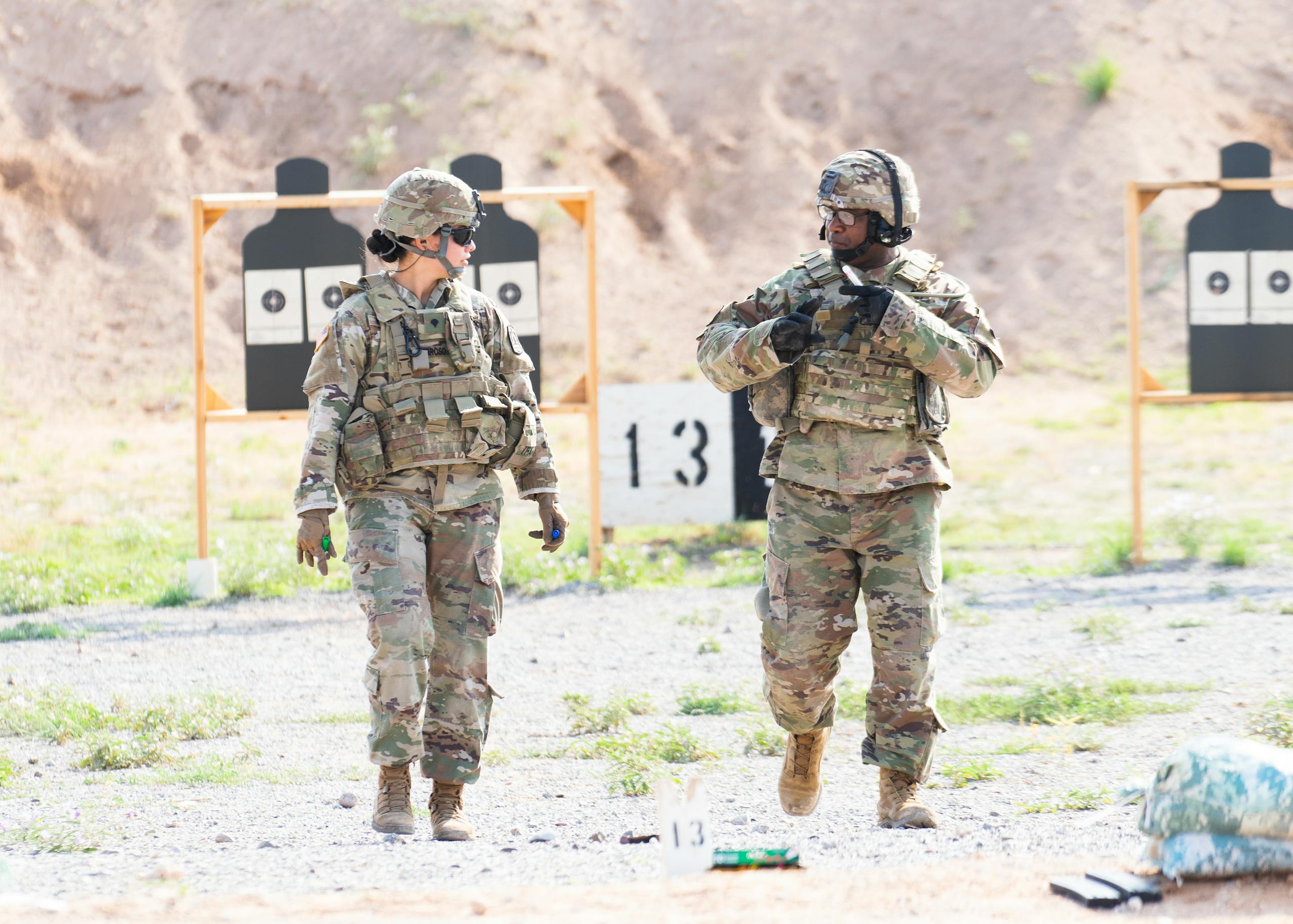 Two soldiers in military uniforms practicing at an outdoor shooting range with targets visible.