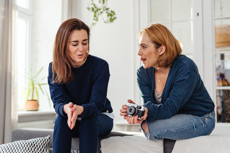 Two women in casual clothes sit on a couch, chatting and smiling warmly indoors.