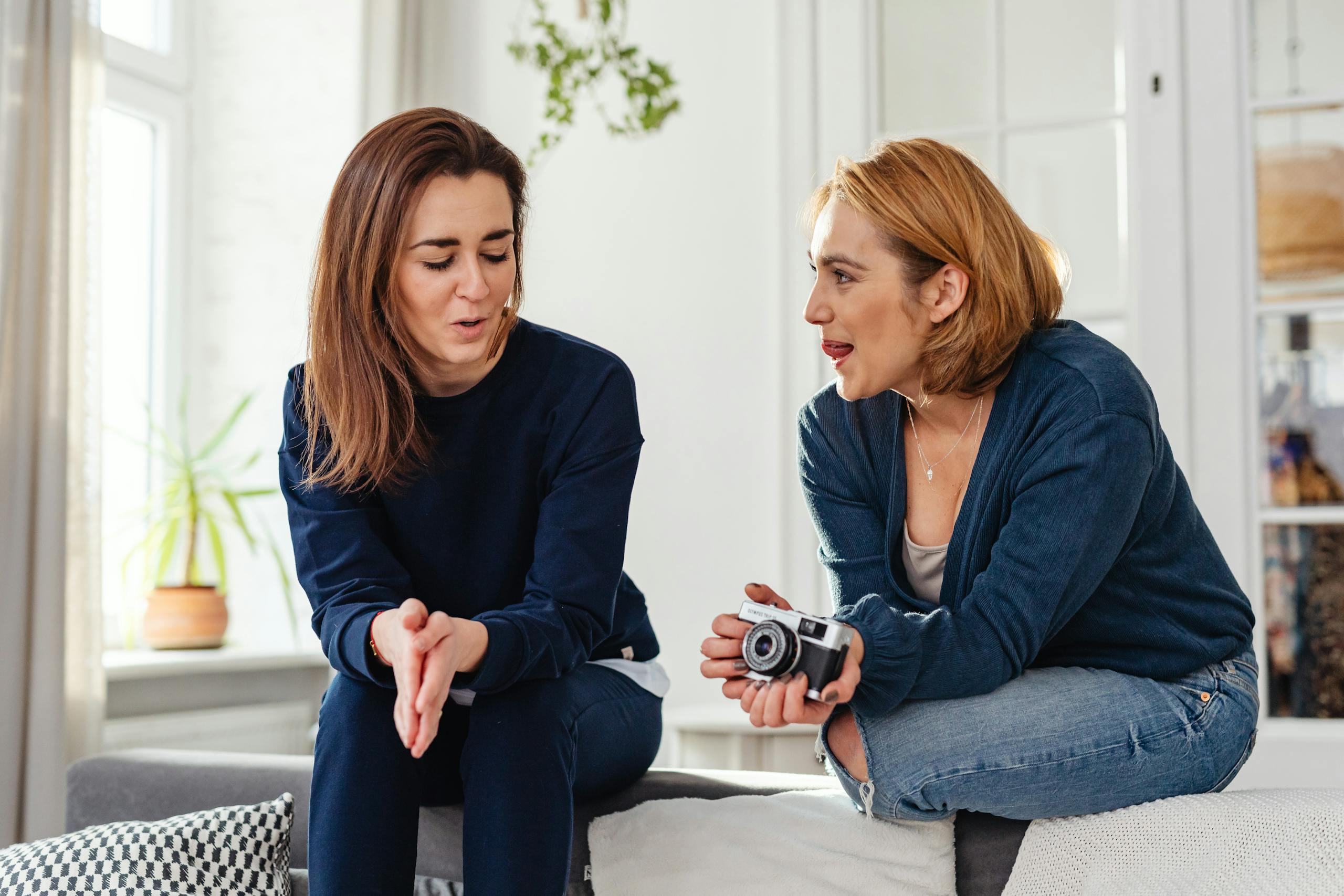 Two women in casual clothes sit on a couch, chatting and smiling warmly indoors.