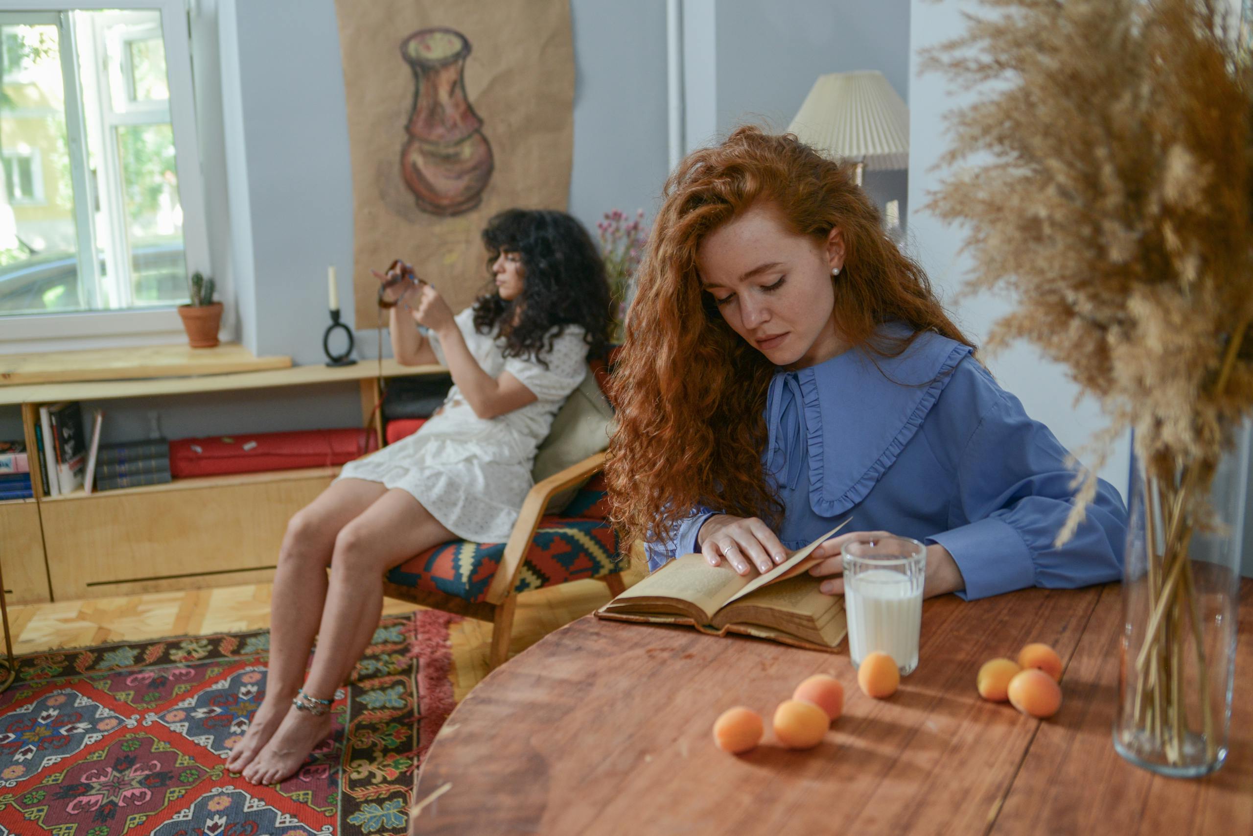 Two women relaxing at home; one reading with a glass of milk, the other using a phone.