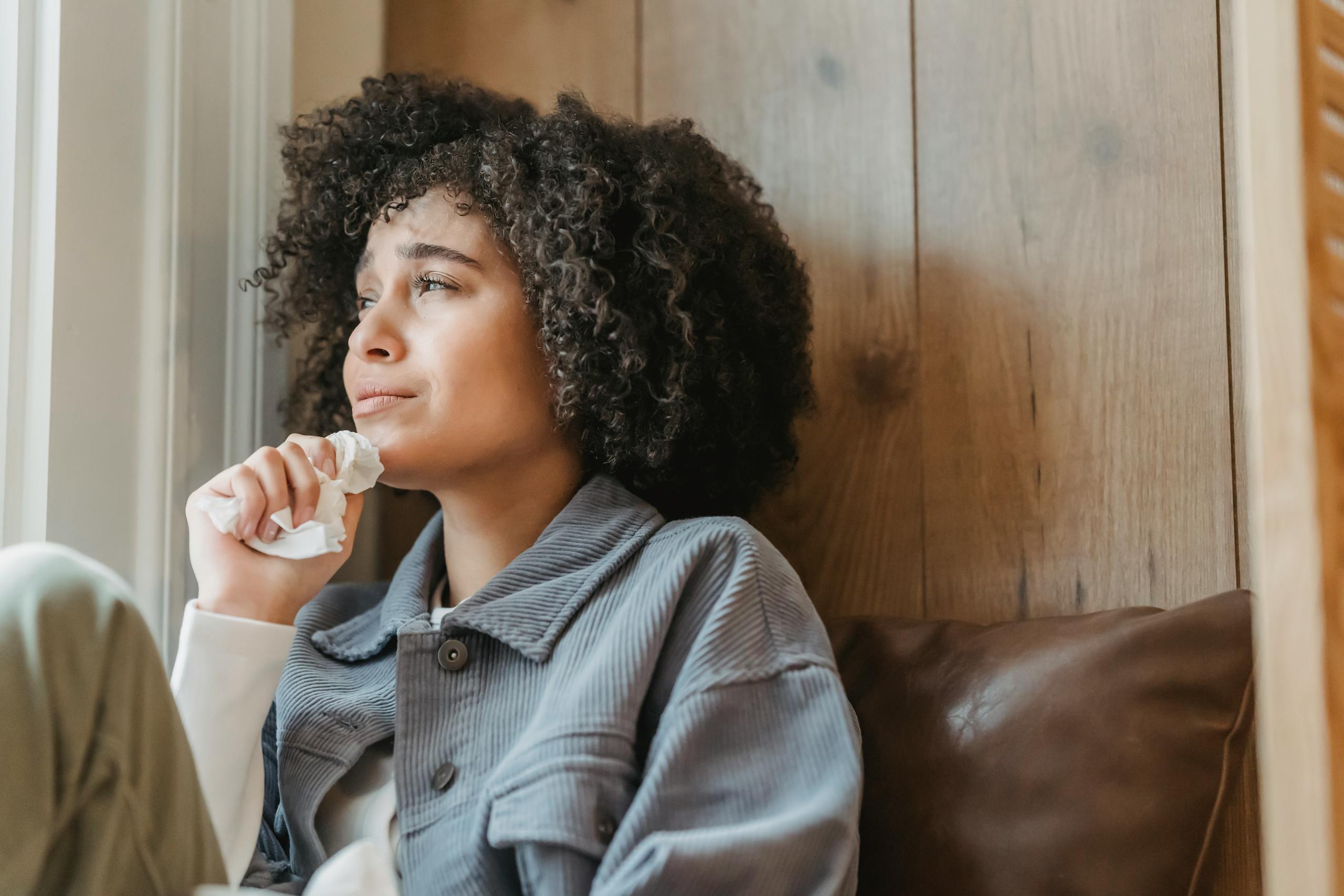 Unhappy young African American female with dark curly hair in casual clothes crying and looking away while sitting near window at home