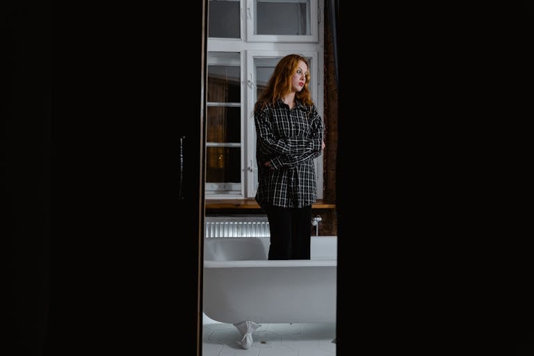 Woman in a flannel shirt standing by a bathtub, reflecting alone.