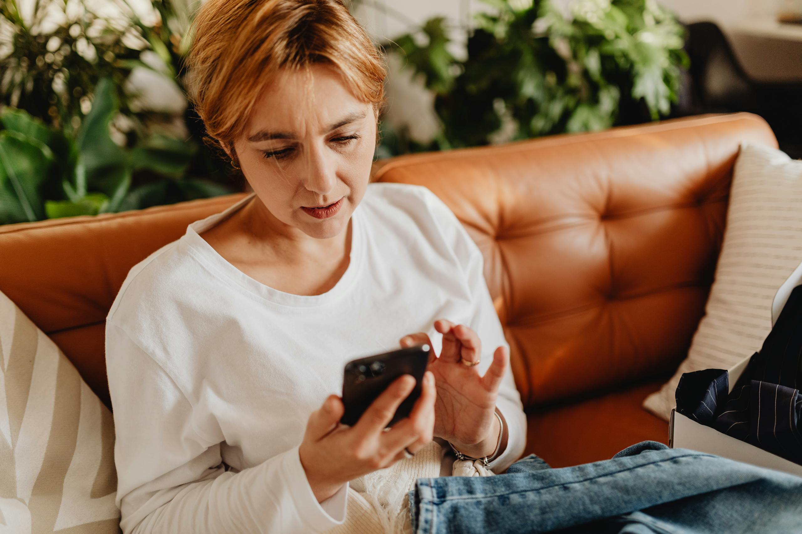 Woman in casual attire using a smartphone on a cozy sofa surrounded by plants.