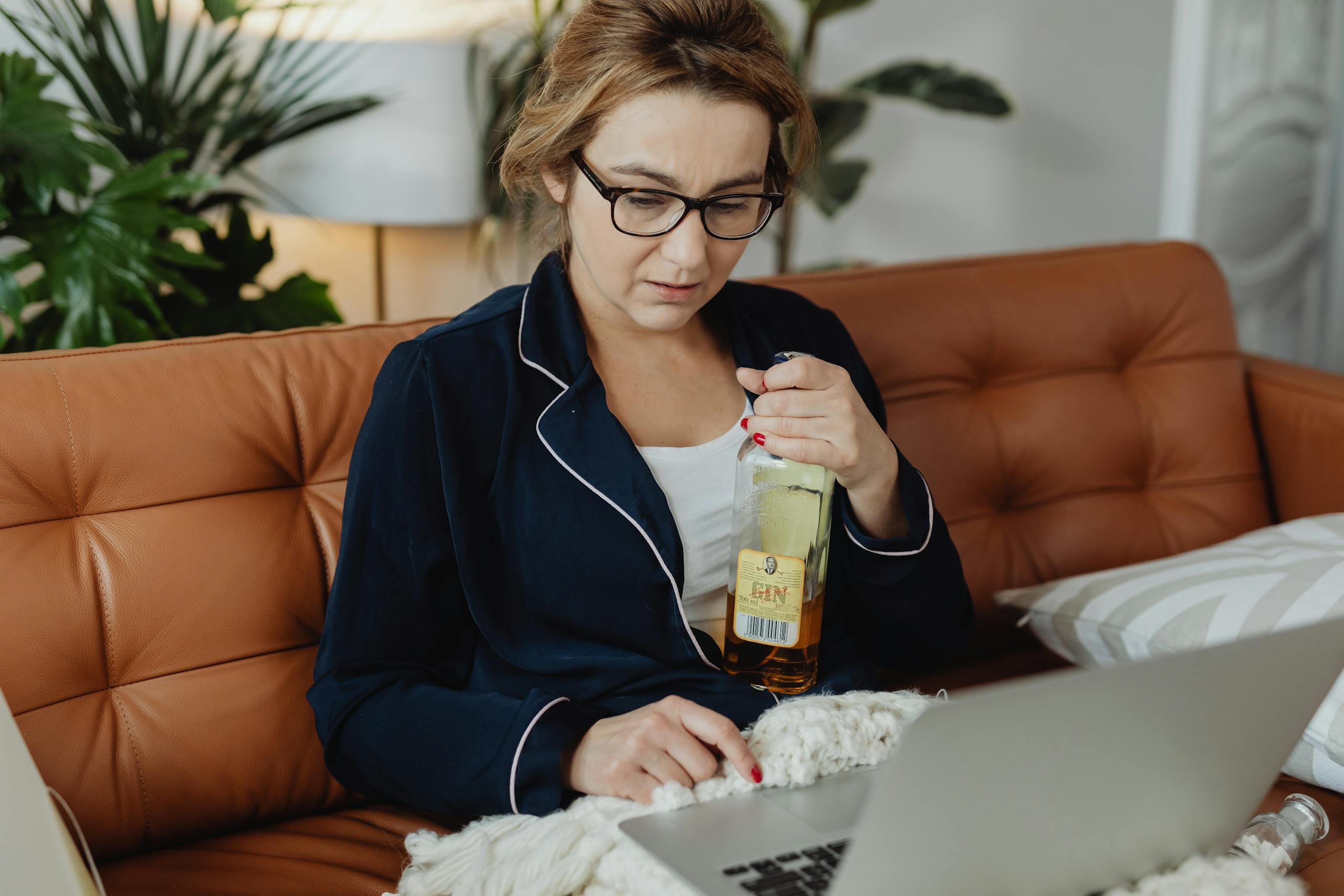 Woman in pajamas holding a bottle of alcohol while working on a laptop, sitting on a sofa at home.