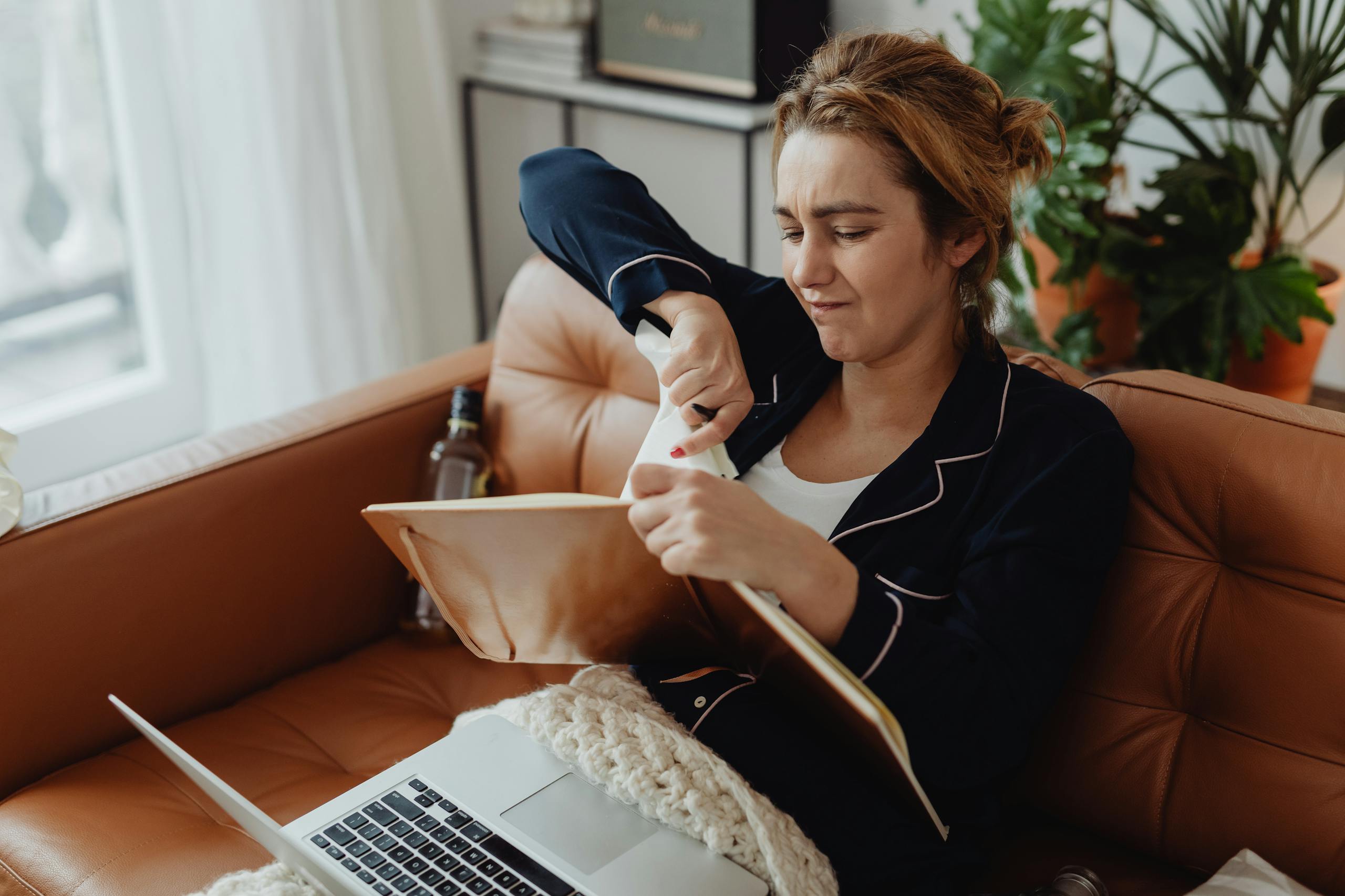 Woman in pajamas tearing paper on a sofa, expressing frustration. Indoors setting with laptop nearby.