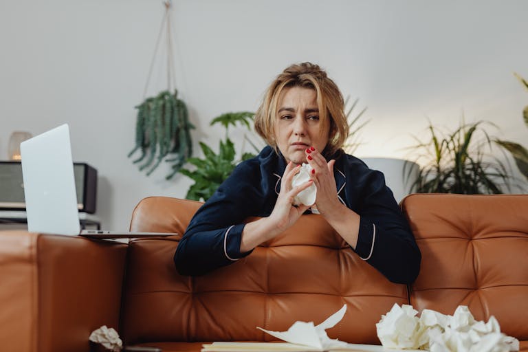 Woman sitting indoors on a brown sofa, holding tissue paper with a sad expression.