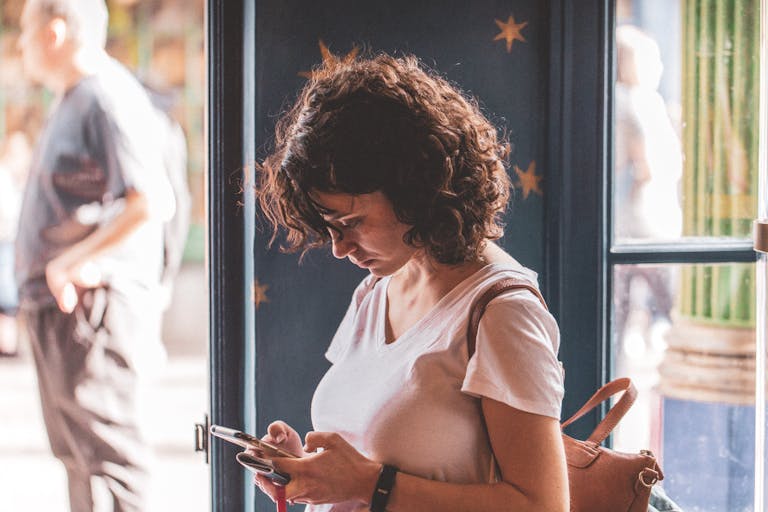 Woman with curly hair using smartphone by a sunlit window, wearing a white t-shirt.