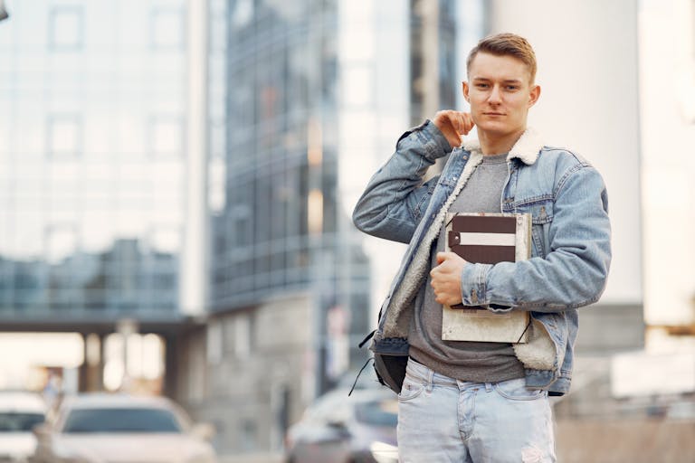 Young adult male student wearing denim jacket, standing confidently outdoors with notebooks.