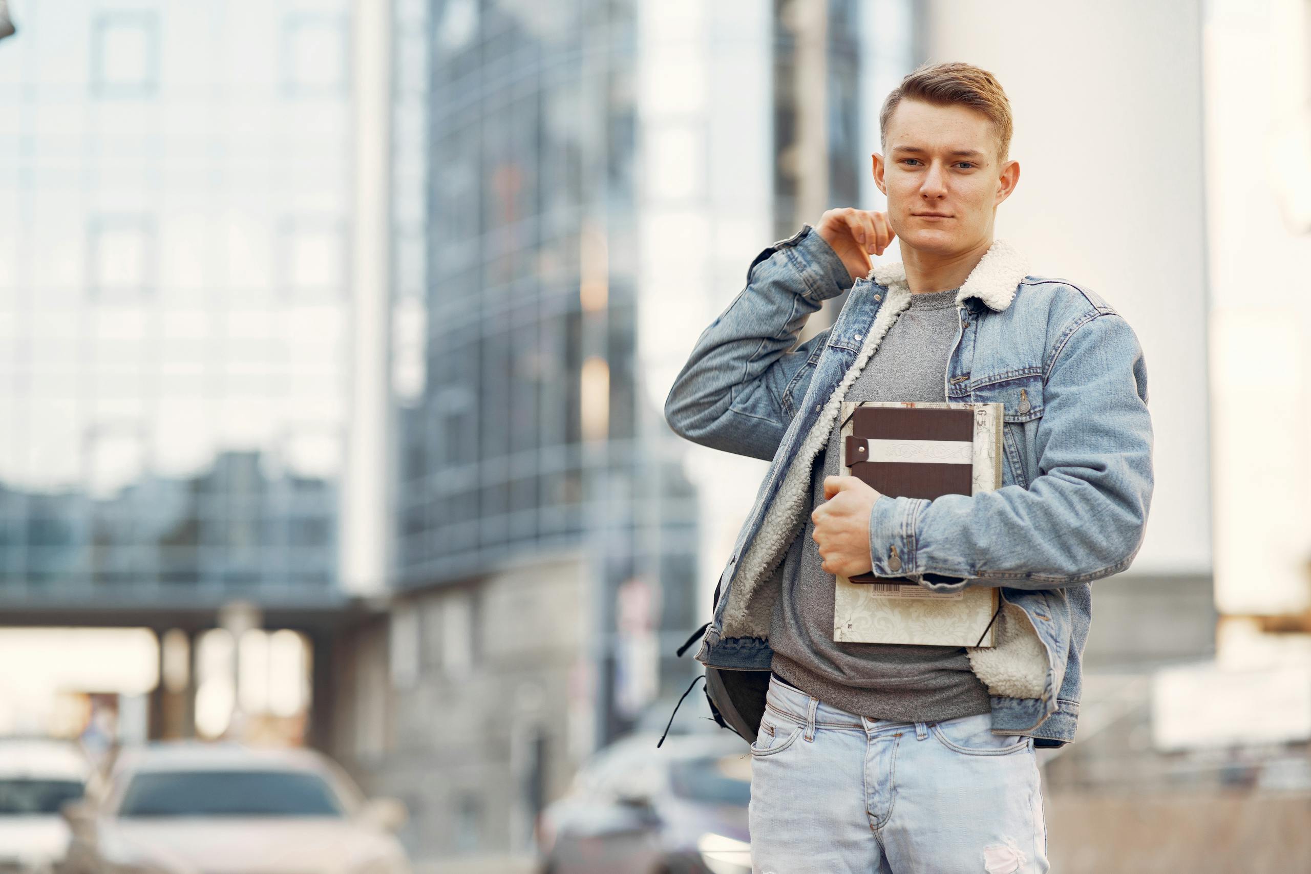 Young adult male student wearing denim jacket, standing confidently outdoors with notebooks.