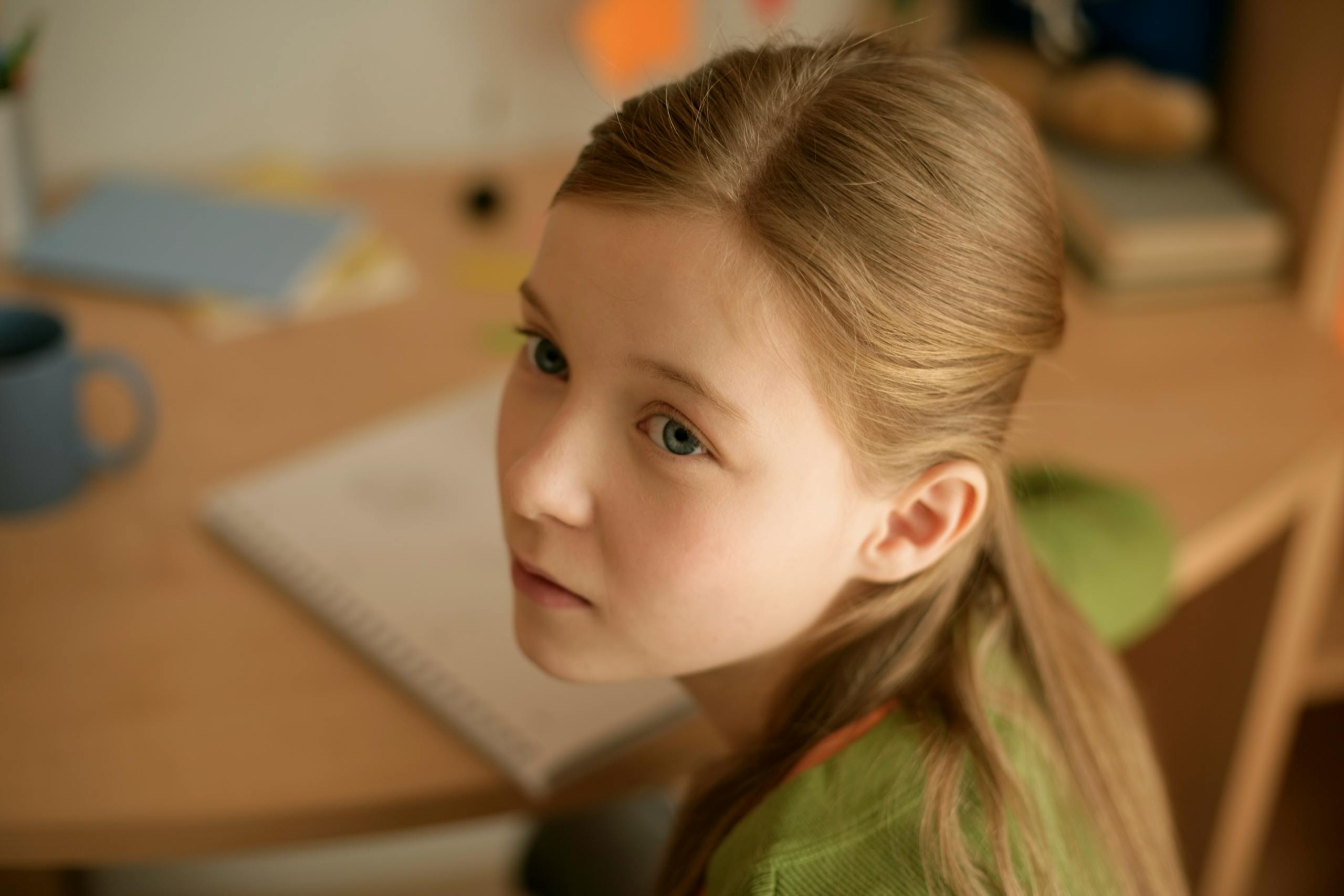 Young girl sitting at a desk in a cozy room, appearing thoughtful and pensive.