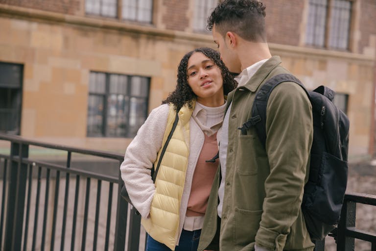 Young Latin American couple wearing warm clothes and backpacks while standing in street near metal railing and building in town and talking in daytime