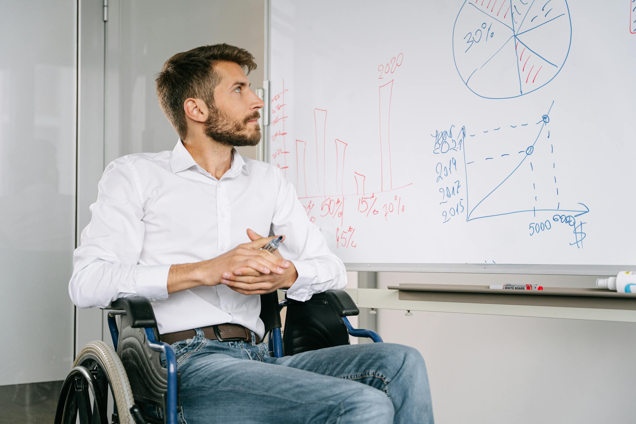 Young man in wheelchair analyzing charts on whiteboard in office setting.