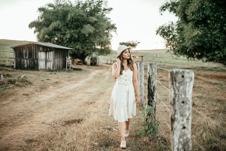 Young woman in white dress walking along a rustic path by a fence, exuding a serene and fashionable vibe.
