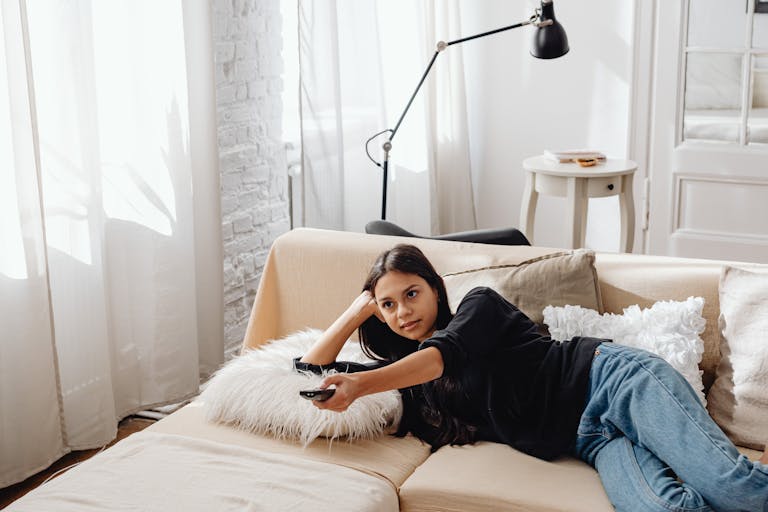 Young woman lounging on a cozy sofa, holding a remote control while relaxing at home.