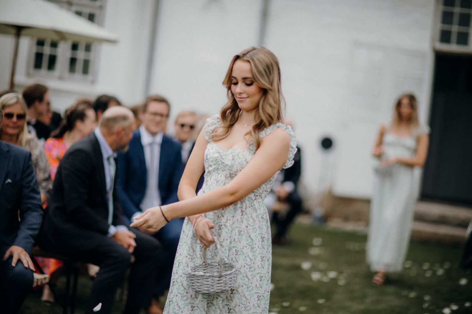 A young woman in a floral dress scattering petals at an outdoor wedding.