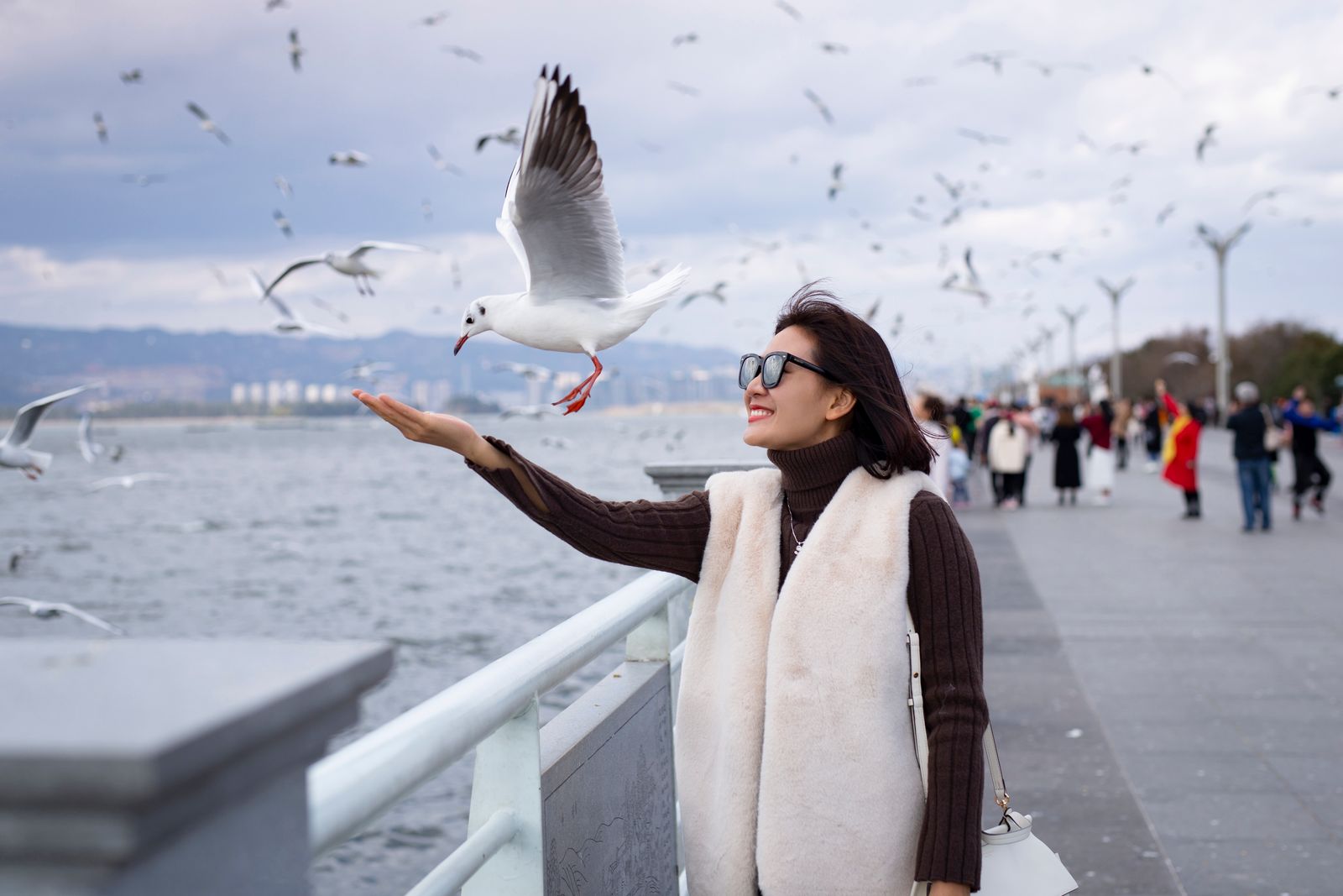Woman joyfully feeds a seagull by the waterfront, surrounded by a flock, under a cloudy sky.