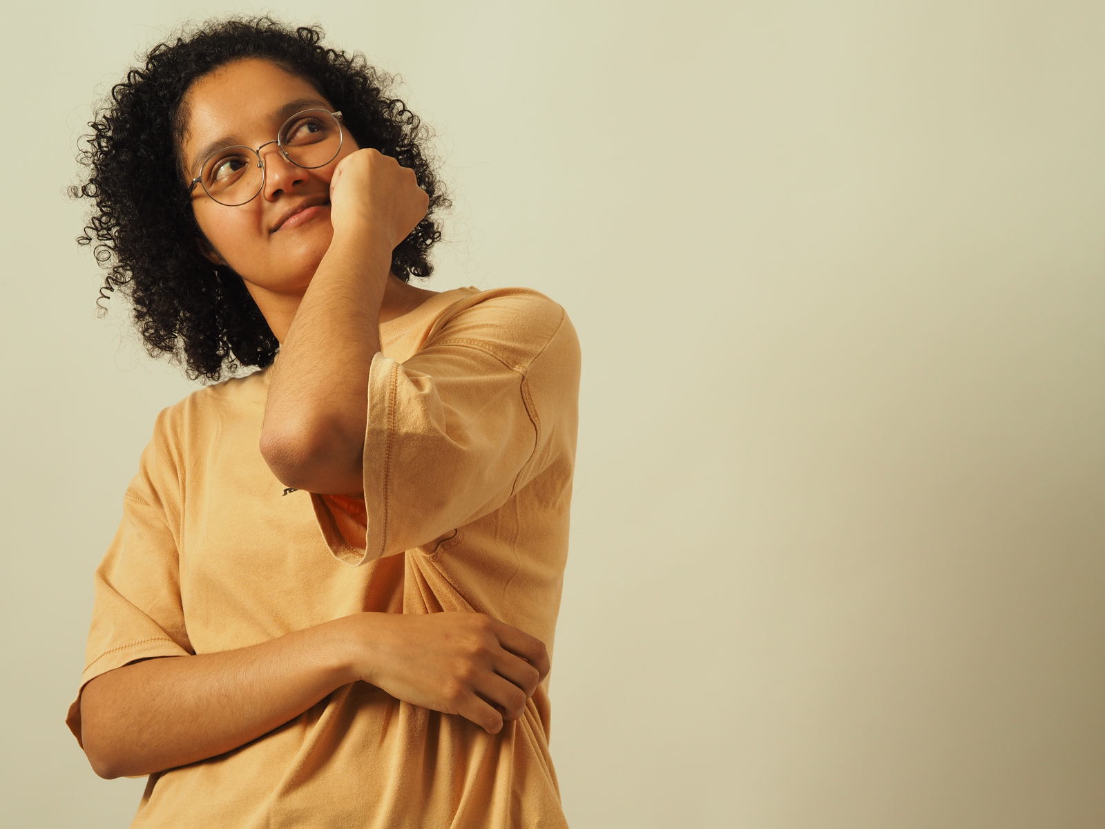Thoughtful person with curly hair and glasses in a casual pose against a neutral background.