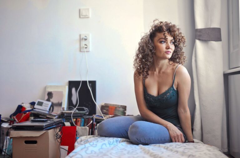 Curly-haired woman sitting pensively on a bed in a cluttered room, looking thoughtful.