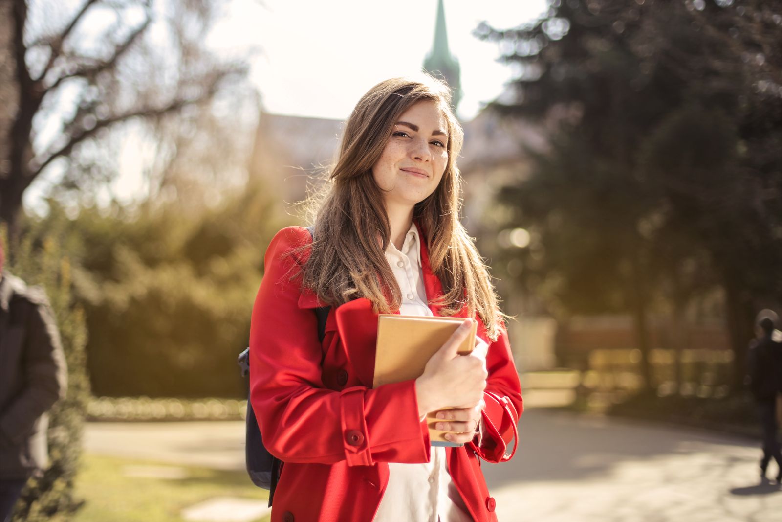 Young woman in a red coat smiling while holding books outdoors on a sunny day.